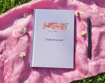 May include: A white gratitude journal with the text "Just Zen It" and "Self Care" in pink lettering. The journal is on a pink blanket with white flowers and a black pen.