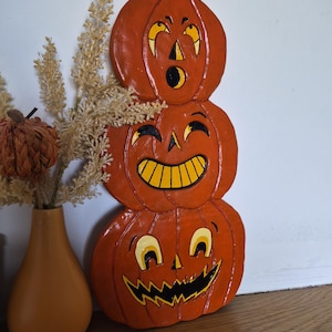 May include: A vertical stack of three orange pumpkins with painted faces. The top pumpkin has a surprised expression, the middle has a wide grin, and the bottom has a jagged, toothy smile. Each pumpkin features black and yellow painted details.
