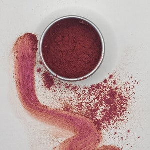 May include: A small metal container filled with a deep red powder. The powder has been spilled out of the container, creating a light red streak on the white surface.