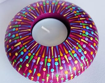 May include: A round wooden candle holder with a tealight well in the center. The holder is painted in a vibrant magenta color with a colorful mandala design featuring dots and lines in shades of yellow, orange, green, blue, and purple.
