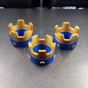 May include: Three 3D-printed crown-shaped objects. Each crown has a gold-colored top and a blue base. The base features gold diamond-shaped accents. The crowns are arranged on a gray surface.