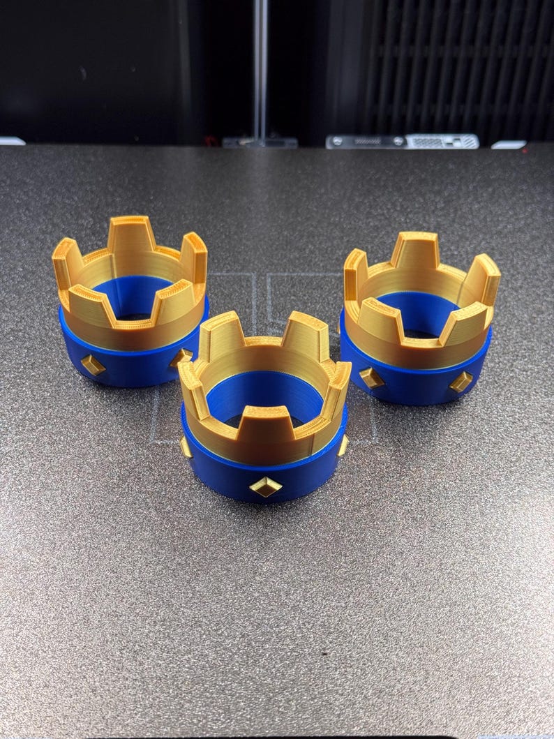 May include: Three 3D-printed crown-shaped objects. Each crown has a gold-colored top and a blue base. The base features gold diamond-shaped accents. The crowns are arranged on a gray surface.
