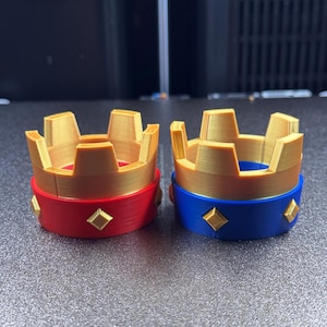 May include: Two 3D-printed crown models. One crown has a red base with gold accents and diamond shapes. The other crown has a blue base with gold accents and diamond shapes. The crowns are on a gray surface.