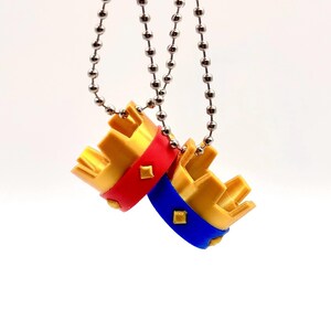May include: Two miniature gold crowns with red and blue bands, each featuring a small diamond-shaped accent. They are suspended from silver ball chains against a white backdrop.