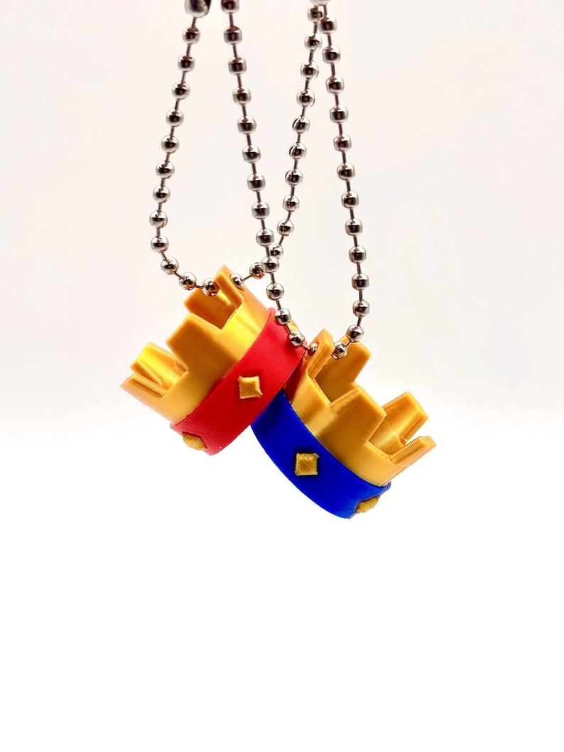 May include: Two miniature gold crowns with red and blue bands, each featuring a small diamond-shaped accent. They are suspended from silver ball chains against a white backdrop.