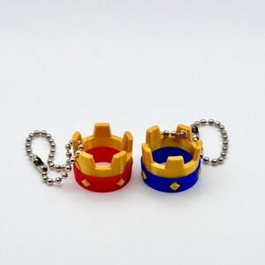 May include: Two miniature crown keyrings. One is red with gold accents, the other is blue with gold accents. Both have a silver chain attached. The crowns have a diamond-shaped design on the base.