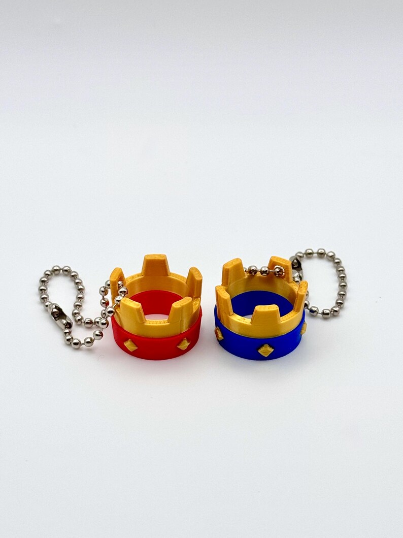 May include: Two miniature crown keyrings. One is red with gold accents, the other is blue with gold accents. Both have a silver chain attached. The crowns have a diamond-shaped design on the base.