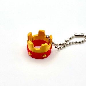 May include: A miniature crown keyring in red and yellow, with a silver-coloured ball chain. The crown has a circular base and a series of upright rectangular shapes on top. The keyring is a small decorative item.