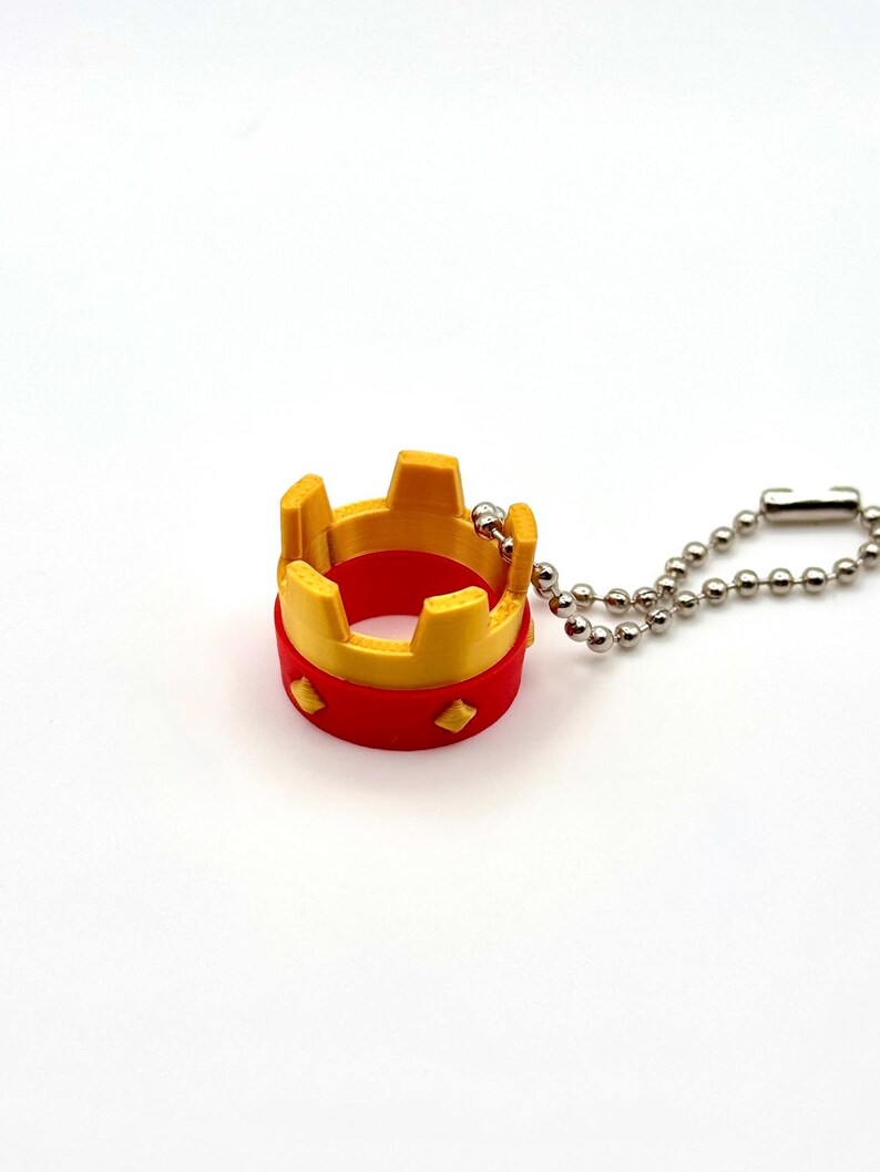 May include: A miniature crown keyring in red and yellow, with a silver-coloured ball chain. The crown has a circular base and a series of upright rectangular shapes on top. The keyring is a small decorative item.