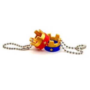 May include: Two miniature crown keyrings. One crown is red and gold, the other is blue and gold. Each crown is attached to a silver ball chain. The keyrings have a royal theme.