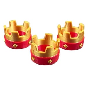 May include: Three miniature gold and red crowns. The crowns have a gold top with a crenelated design and a red base with diamond-shaped accents. The crowns are likely decorative or for a craft project.