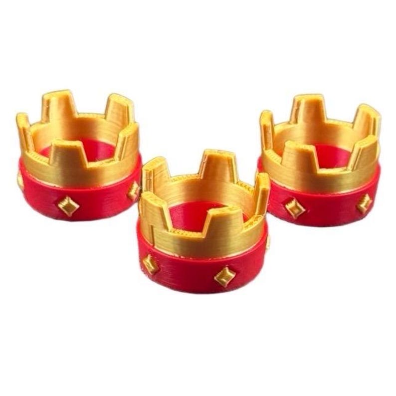May include: Three miniature gold and red crowns. The crowns have a gold top with a crenelated design and a red base with diamond-shaped accents. The crowns are likely decorative or for a craft project.