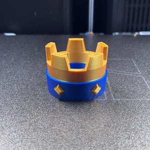 May include: A 3D-printed crown in gold and blue. The crown has a gold top with five points and a blue base. The base is decorated with four gold diamond shapes. The crown is likely a toy or decorative item.
