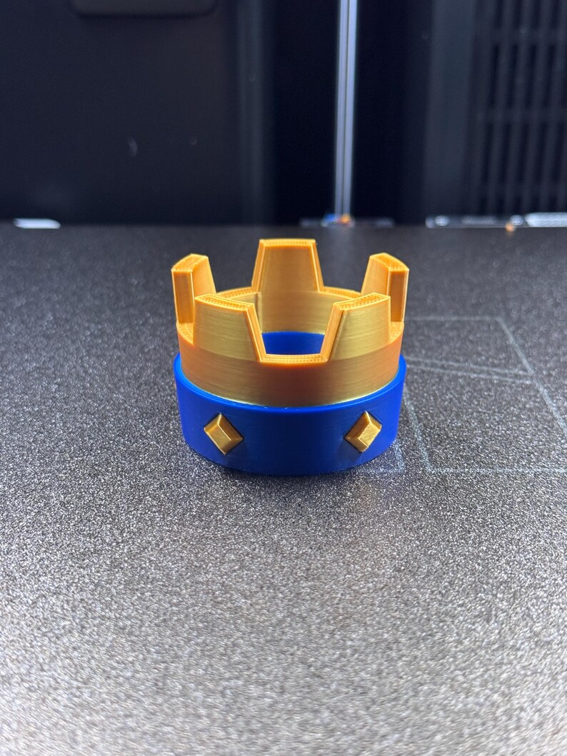 May include: A 3D-printed crown in gold and blue. The crown has a gold top with five points and a blue base. The base is decorated with four gold diamond shapes. The crown is likely a toy or decorative item.