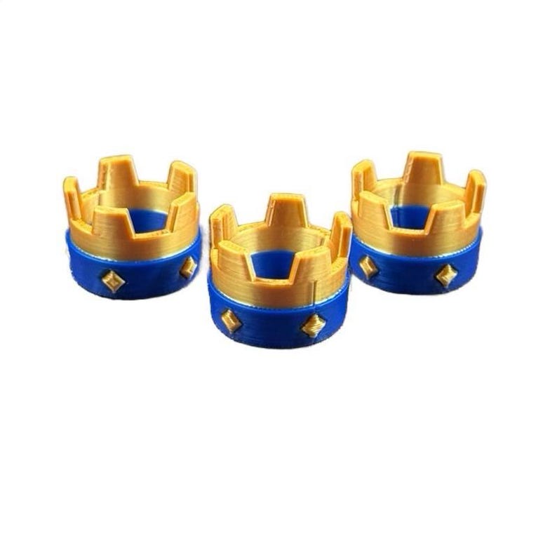 May include: Three miniature gold and blue crown-shaped objects. The crowns have a gold top with a blue base, each adorned with diamond-shaped accents. The objects are arranged on a white background.