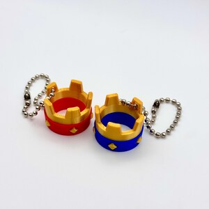 May include: Two miniature crown keyrings. One is red with gold accents, the other is blue with gold accents. Both have a diamond pattern and are attached to a silver ball chain.