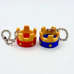 May include: Two miniature crown keyrings. One has a red base with gold accents, and the other has a blue base with gold accents. Both have a diamond pattern on the base and are attached to a silver ball chain.