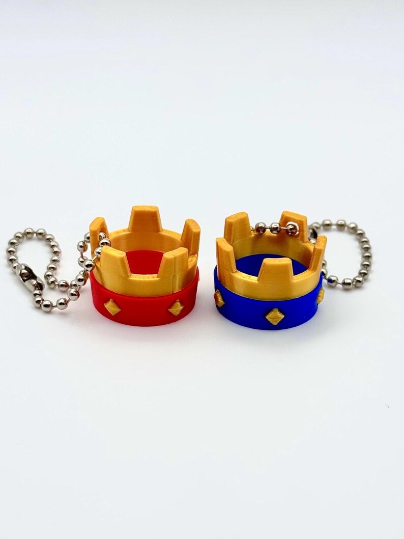 May include: Two miniature crown keyrings. One has a red base with gold accents, and the other has a blue base with gold accents. Both have a diamond pattern on the base and are attached to a silver ball chain.