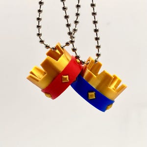 May include: Two small, gold-coloured crown pendants with red and blue bands, each adorned with a diamond-shaped accent. The crowns hang from silver ball chains against a white background. The pendants are likely jewellery or decorative items.