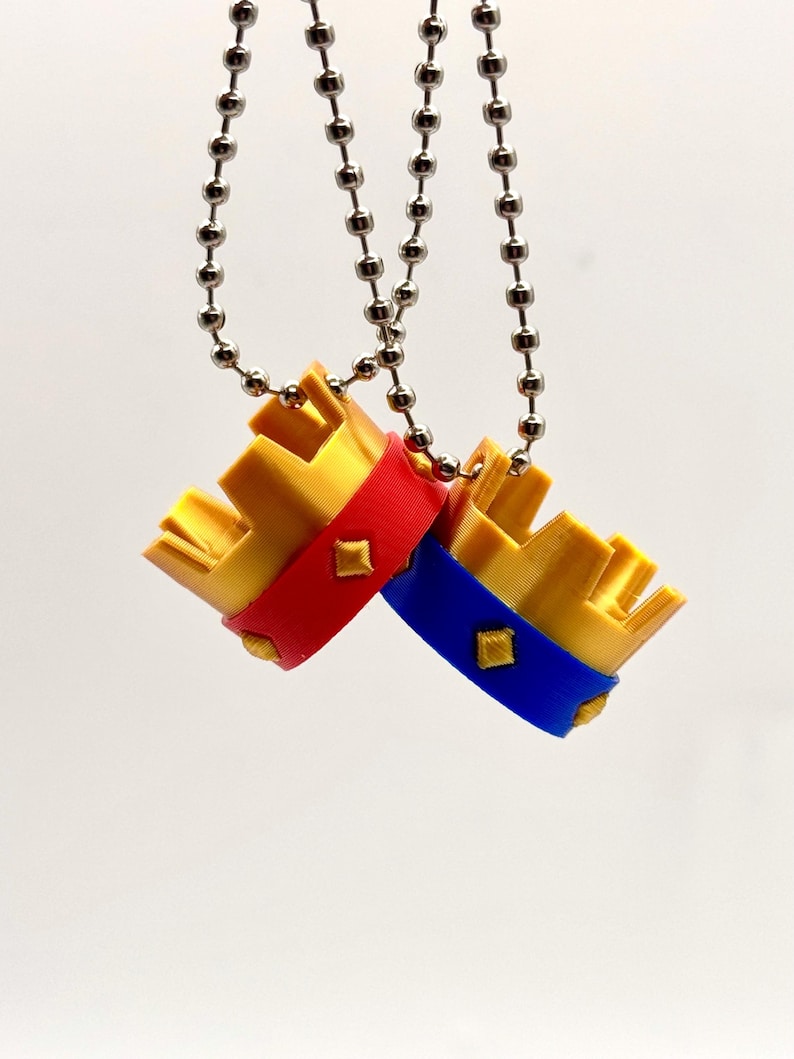 May include: Two small, gold-coloured crown pendants with red and blue bands, each adorned with a diamond-shaped accent. The crowns hang from silver ball chains against a white background. The pendants are likely jewellery or decorative items.