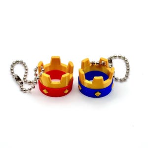 May include: Two miniature crown keyrings. One is red and gold, the other is blue and gold. Each crown has a diamond-shaped accent and is attached to a silver ball chain. The keyrings are set against a white background.