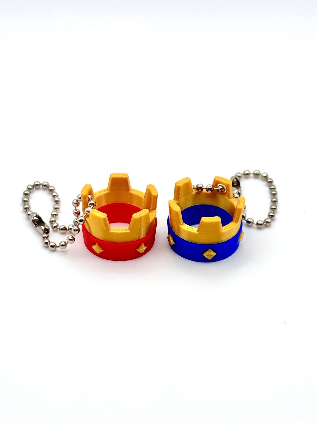 May include: Two miniature crown keyrings. One is red and gold, the other is blue and gold. Each crown has a diamond-shaped accent and is attached to a silver ball chain. The keyrings are set against a white background.
