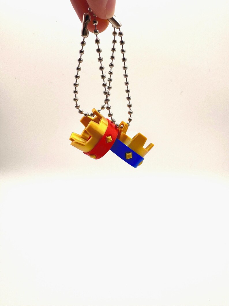May include: Two miniature crown keyrings. One crown has a red band, the other has a blue band. Both crowns are gold with silver chain attachments. The keyrings are suspended against a white background.