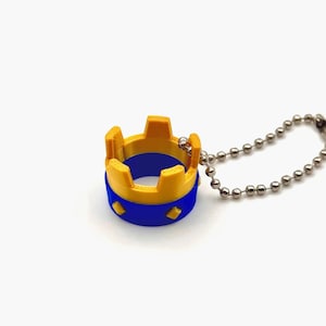May include: A miniature crown-shaped pendant on a ball chain. The crown features a yellow top and a blue band with diamond shapes. The chain is made of small, silver-coloured balls. The design is simple and geometric.