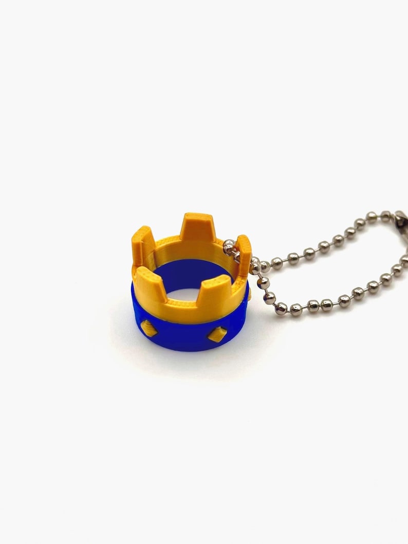 May include: A miniature crown-shaped pendant on a ball chain. The crown features a yellow top and a blue band with diamond shapes. The chain is made of small, silver-coloured balls. The design is simple and geometric.