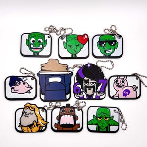 May include: A collection of cartoon character dog tag keychains. Each tag features a unique character design with a white background and a silver chain. The characters include green aliens, pigs, and other stylized figures.