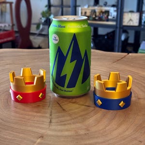 May include: A green can of Alpine Blast Prebiotic Soda with a blue mountain design, next to two decorative crown-shaped objects. One crown is red and gold, the other is blue and gold. The soda can reads "5g Sugar" and "Back, but Better."