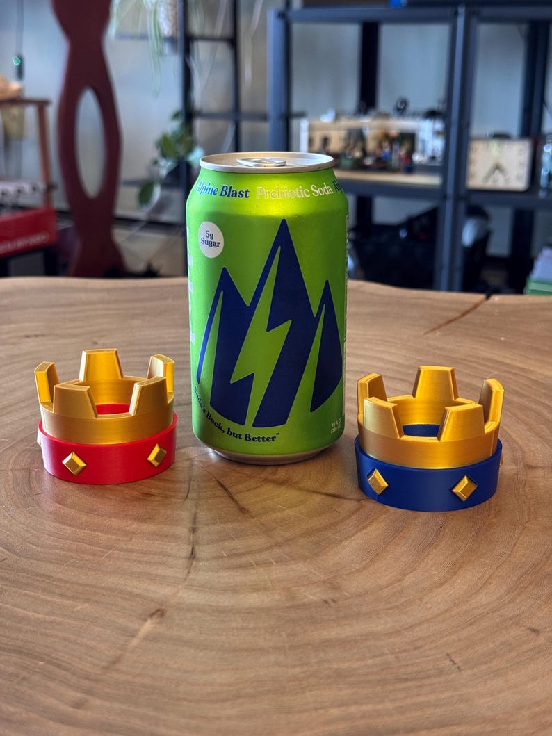 May include: A green can of Alpine Blast Prebiotic Soda with a blue mountain design, next to two decorative crown-shaped objects. One crown is red and gold, the other is blue and gold. The soda can reads "5g Sugar" and "Back, but Better."