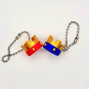 May include: Two miniature gold-coloured crown keychains. One crown has a red band with diamond-shaped accents, the other has a blue band. Both have a silver ball chain attached.