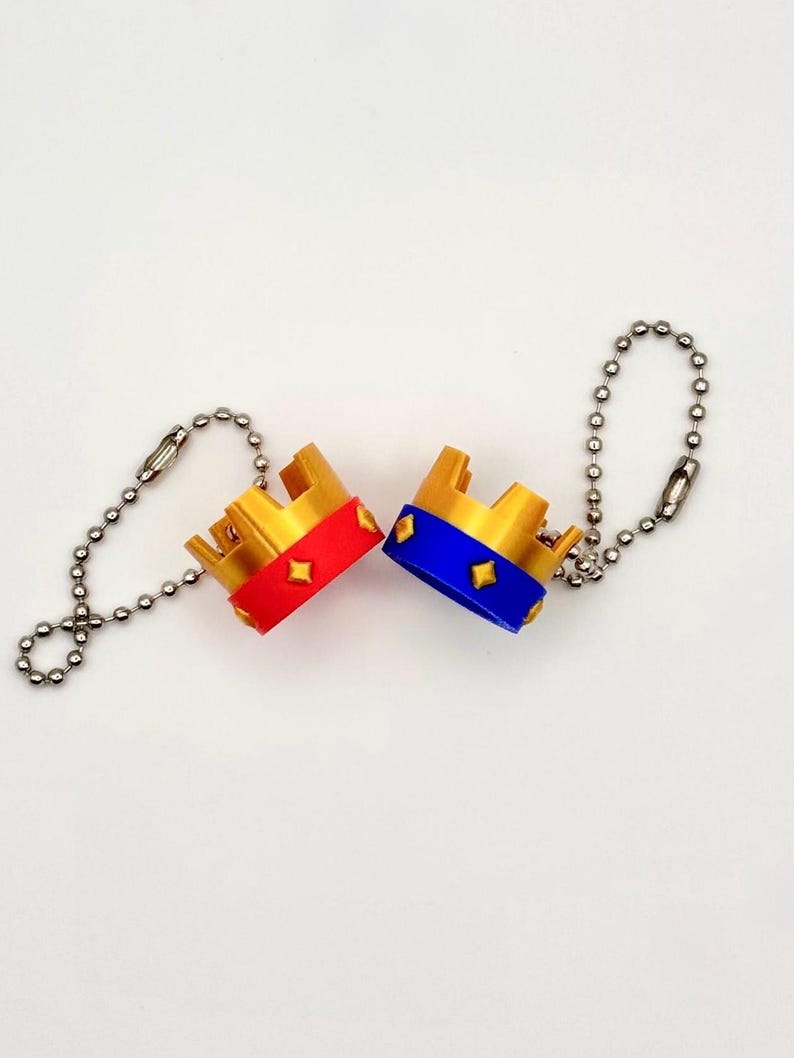 May include: Two miniature gold-coloured crown keychains. One crown has a red band with diamond-shaped accents, the other has a blue band. Both have a silver ball chain attached.