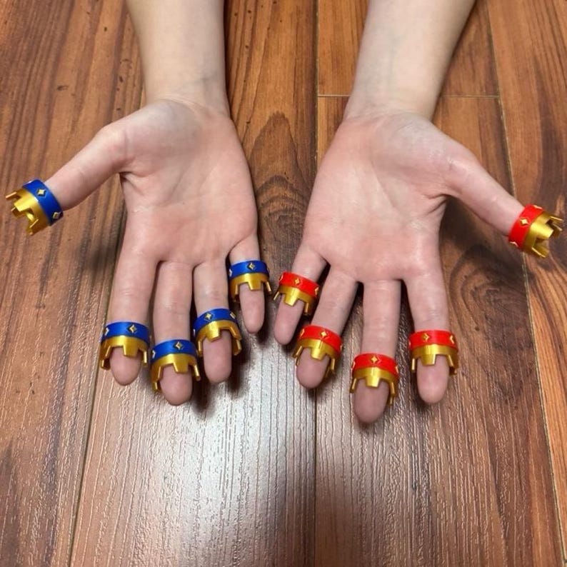 May include: A collection of finger rings in gold, blue, and red. The rings feature a crown design. The blue rings have a gold crown and a blue band with a gold star. The red rings have a gold crown and a red band.
