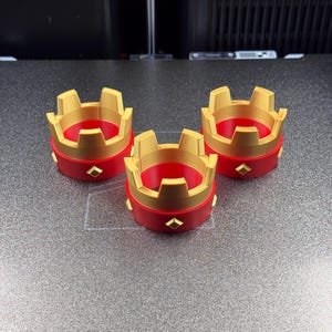 May include: Three red and gold crown-shaped objects with a diamond-shaped accent. The crowns have a gold top and a red base. The objects are likely decorative or part of a larger set.