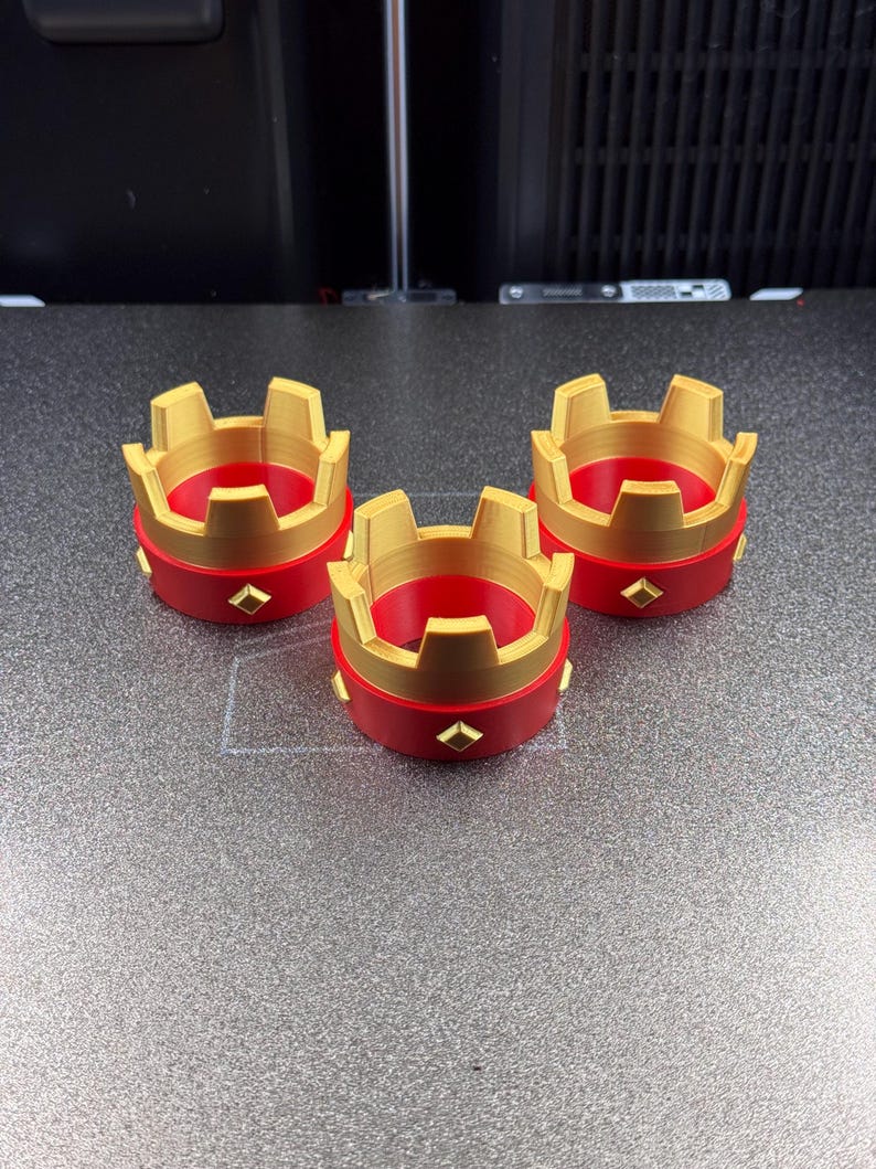 May include: Three red and gold crown-shaped objects with a diamond-shaped accent. The crowns have a gold top and a red base. The objects are likely decorative or part of a larger set.