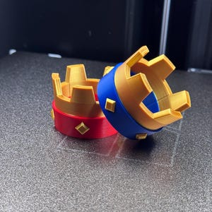 May include: Two small, decorative crowns. One crown has a red band with gold diamond accents and a gold crown top. The other crown has a blue band with gold diamond accents and a gold crown top. The crowns are made of plastic.