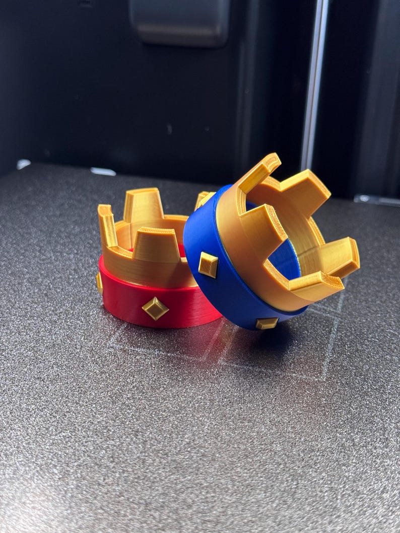 May include: Two small, decorative crowns. One crown has a red band with gold diamond accents and a gold crown top. The other crown has a blue band with gold diamond accents and a gold crown top. The crowns are made of plastic.