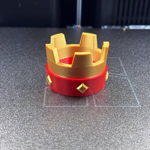 May include: A 3D-printed crown with a gold-colored top and a red base. The crown features a series of rectangular cutouts and is adorned with gold-colored diamond-shaped accents around the red base. The object is likely a decorative item or toy.