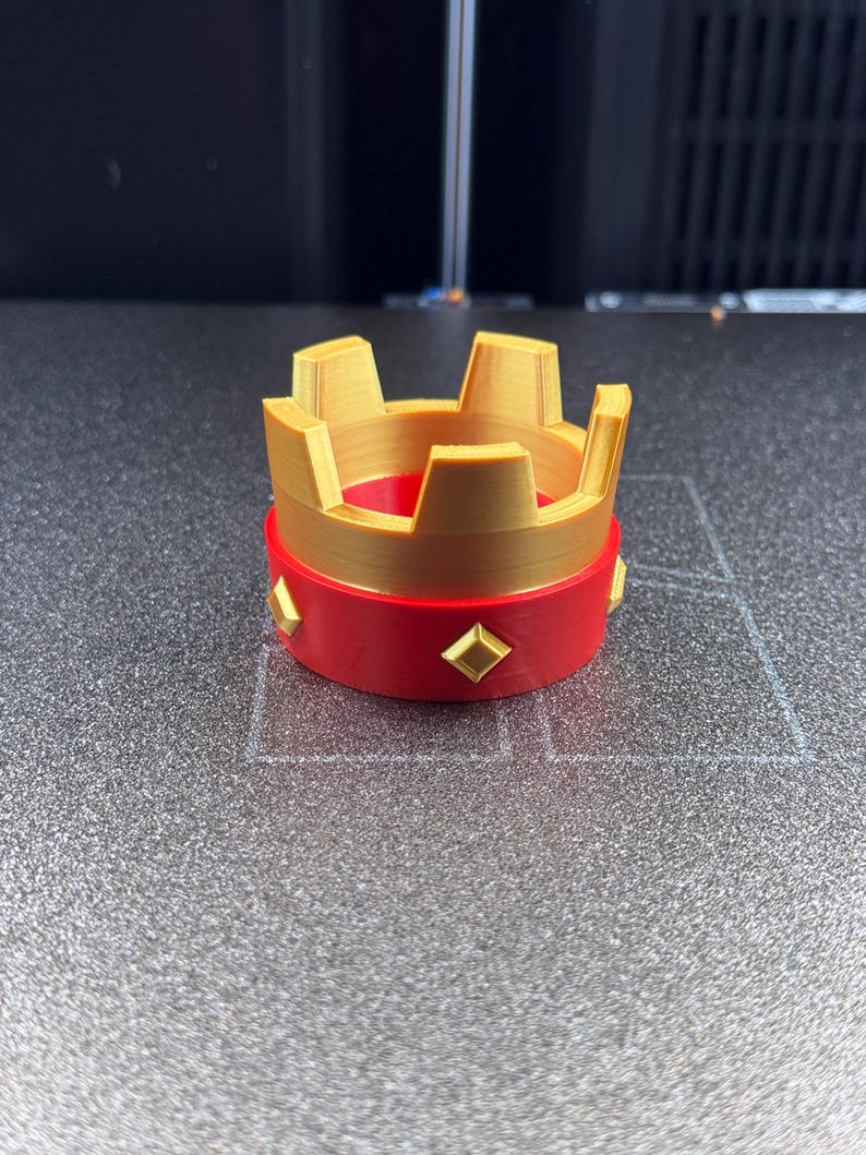 May include: A 3D-printed crown with a gold-colored top and a red base. The crown features a series of rectangular cutouts and is adorned with gold-colored diamond-shaped accents around the red base. The object is likely a decorative item or toy.