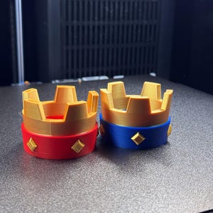 May include: Two 3D-printed crown-shaped objects. One has a red base with gold accents, and the other has a blue base with gold accents. Both have a gold crown top. The crowns are likely decorative or for a craft project.
