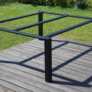 Può includere: Struttura di tavolo in metallo nero con piano rettangolare e quattro gambe quadrate. Il telaio è posizionato su un ponte di legno, con un prato verde sullo sfondo. Progettato per uso esterno.