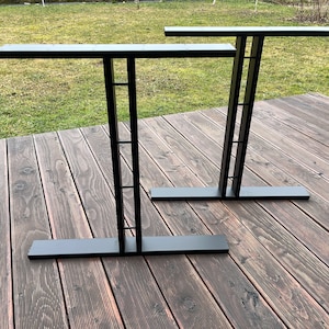 May include: Two black metal table legs with a rectangular top and a square design on the vertical supports. The legs are on a wooden deck.
