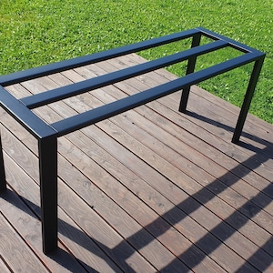 May include: A black metal table frame with a rectangular top and four legs. The frame has three horizontal supports. The table frame is on a wooden deck with green grass in the background.