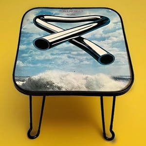 May include: A small square side table with black metal hairpin legs and a tabletop featuring the Tubular Bells album cover art by Mike Oldfield, depicting a bent metallic tube shape against a blue sky with clouds and ocean waves.