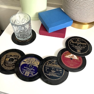 May include: A set of circular coasters designed to resemble vintage vinyl records, featuring classic labels from Brunswick, Decca, Columbia, His Master's Voice, and Parlophone. The black coasters are arranged on a white surface near a glass tumbler.