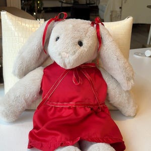 May include: A plush bunny doll with long, floppy ears and a red satin dress. The bunny has a cream-colored body, black eyes, and red ribbons tied in its ears. The dress is trimmed with gold ribbon.
