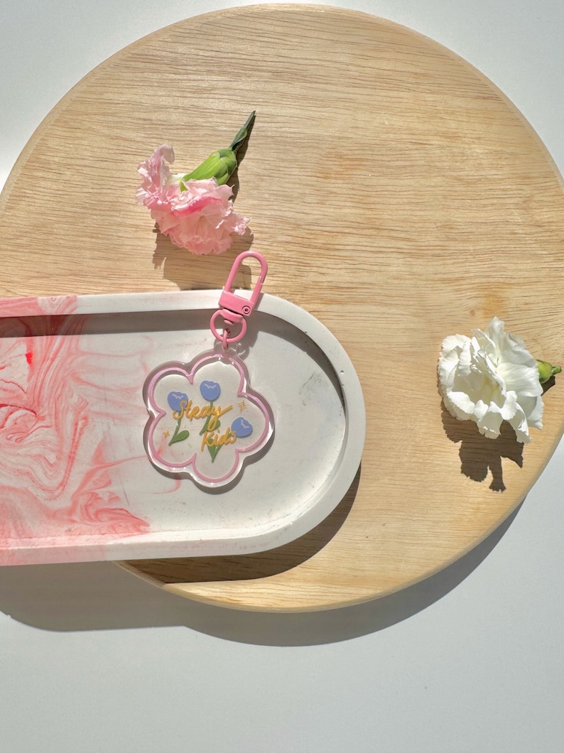 Pu&ograve; includere: Un vassoio ovale rosa e bianco con un portachiavi a forma di fiore rosa con il testo "Sleepy Kids" e fiori blu. Il portachiavi &egrave; sopra il vassoio. Ci sono due garofani rosa e un garofano bianco su una superficie di legno chiaro.