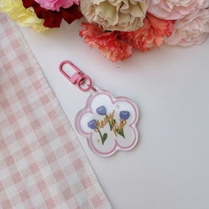 Pu&ograve; includere: Un portachiavi bianco a forma di fiore con un contorno rosa. Il portachiavi ha due fiori blu con steli verdi e una scritta dorata che dice "Sleepy Kids".